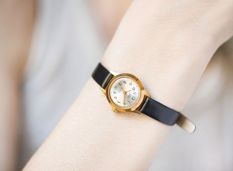 Peut inclure: Une montre-bracelet dor&eacute;e dot&eacute;e d'un bo&icirc;tier rond avec un cadran blanc, des chiffres arabes noirs pour les heures et de fines aiguilles dor&eacute;es. La montre est fix&eacute;e &agrave; un bracelet en cuir noir fin et lisse avec une boucle dor&eacute;e et une couronne.