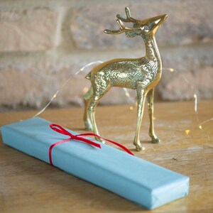 May include: A polished brass deer figurine standing next to a rectangular gift box wrapped in light blue paper and tied with a thin red ribbon. The deer is depicted in a standing position with its head tilted upward, featuring textured metallic details.