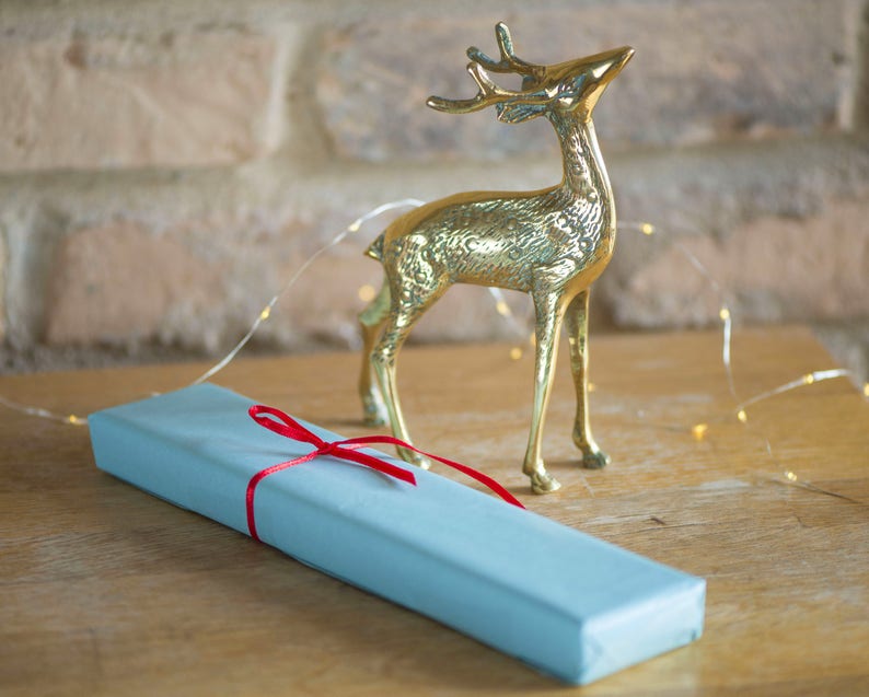 May include: A polished brass deer figurine standing next to a rectangular gift box wrapped in light blue paper and tied with a thin red ribbon. The deer is depicted in a standing position with its head tilted upward, featuring textured metallic details.