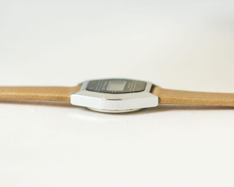 May include: A digital wristwatch featuring a silver-toned metallic casing with a black display screen and a light tan textured strap. The watch is positioned horizontally, showcasing the side profile of the metallic frame and the strap attachment.
