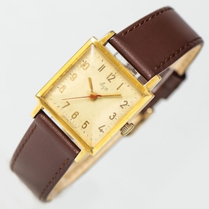 May include: A gold-tone square wristwatch with a white face and black Roman numeral hour markers. The watch has a brown leather band and the text "Слава" on the face.