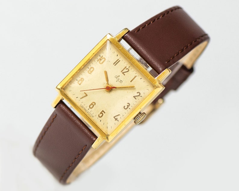 May include: A gold-tone square wristwatch with a white face and black Roman numeral hour markers. The watch has a brown leather band and the text "Слава" on the face.