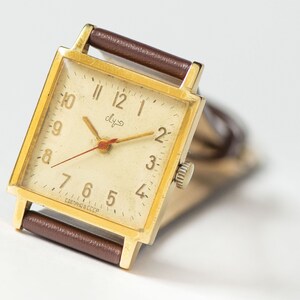May include: A gold-toned square wristwatch with a white face and black Roman numeral hour markers. The watch has a brown leather band and says "СССР" on the face.