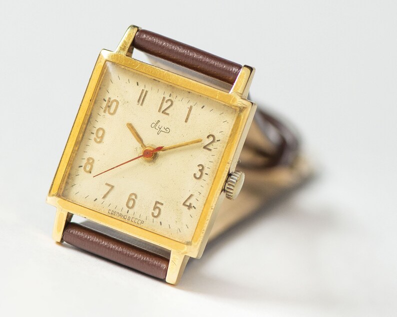 May include: A gold-toned square wristwatch with a white face and black Roman numeral hour markers. The watch has a brown leather band and says "СССР" on the face.
