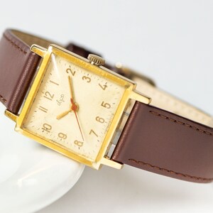 May include: A gold-toned square wristwatch with a brown leather band. The watch face has a white background with black Roman numeral hour markers and black hands. The watch face also has the word "dlya" printed in black.