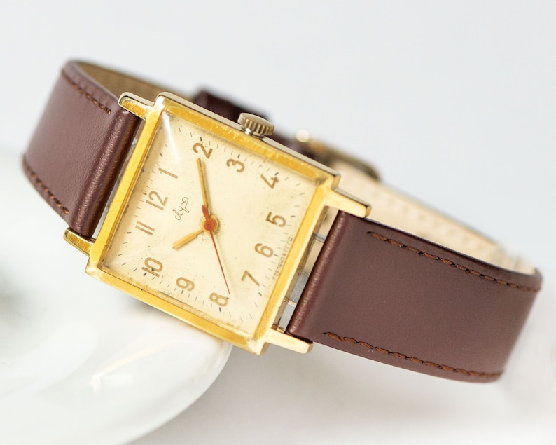 May include: A gold-toned square wristwatch with a brown leather band. The watch face has a white background with black Roman numeral hour markers and black hands. The watch face also has the word "dlya" printed in black.