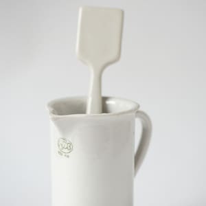 May include: A white ceramic jug with a handle and a white ceramic spatula inside. The jug has a green logo that says "150 ml".