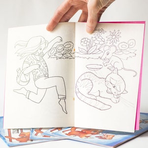 May include: A coloring book with a pink cover and white pages. The book features a girl running with a watering can, a cat sleeping, and a mouse. The book is open to a page with a girl running with a watering can on the left side and a cat sleeping on the right side.