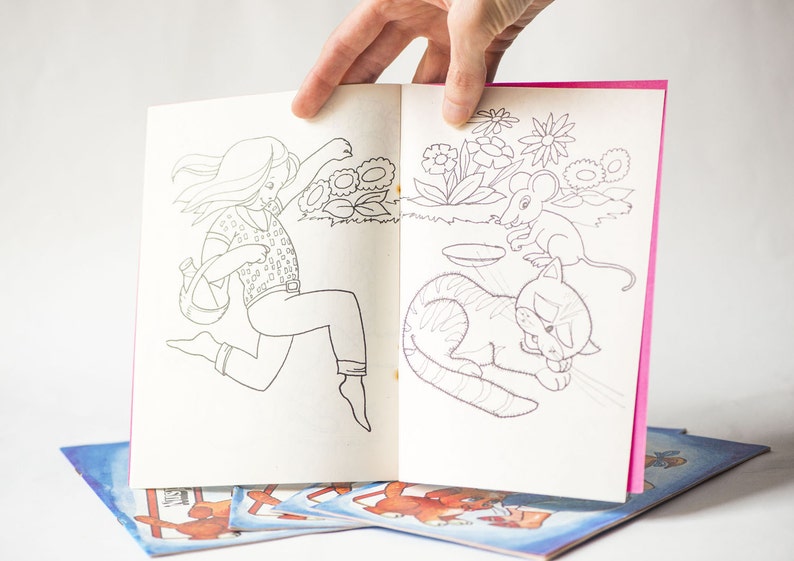 May include: A coloring book with a pink cover and white pages. The book features a girl running with a watering can, a cat sleeping, and a mouse. The book is open to a page with a girl running with a watering can on the left side and a cat sleeping on the right side.