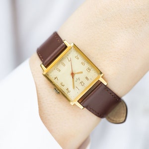 May include: A gold-toned square watch with a brown leather band. The watch face is white with black Roman numeral hour markers and black Arabic numeral minute markers. The watch has a red second hand.