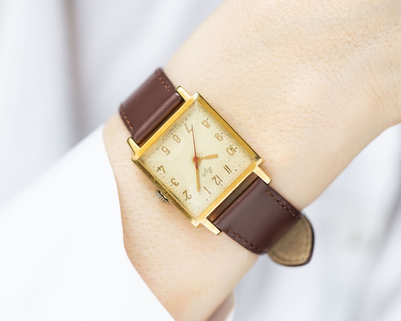 May include: A gold-toned square watch with a brown leather band. The watch face is white with black Roman numeral hour markers and black Arabic numeral minute markers. The watch has a red second hand.