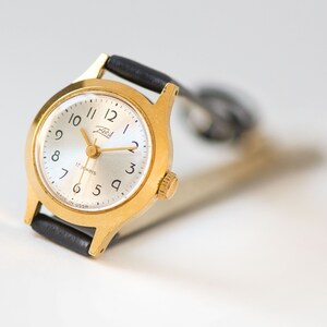 Peut inclure: Une montre-bracelet dor&eacute;e avec un cadran argent&eacute; rond, des chiffres arabes noirs et des aiguilles dor&eacute;es. Le cadran porte l'inscription Zaria et 17 jewels, avec Made in USSR imprim&eacute; en bas. Elle comprend un bracelet en cuir noir avec boucle.