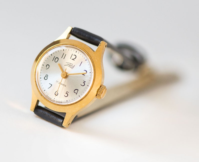 Peut inclure: Une montre-bracelet dor&eacute;e avec un cadran argent&eacute; rond, des chiffres arabes noirs et des aiguilles dor&eacute;es. Le cadran porte l'inscription Zaria et 17 jewels, avec Made in USSR imprim&eacute; en bas. Elle comprend un bracelet en cuir noir avec boucle.