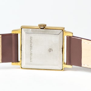 May include: A gold-tone square wristwatch with a brown leather band. The watch face is silver and has the text "ПРОТИВОУДАРНЫЕ" printed on it.