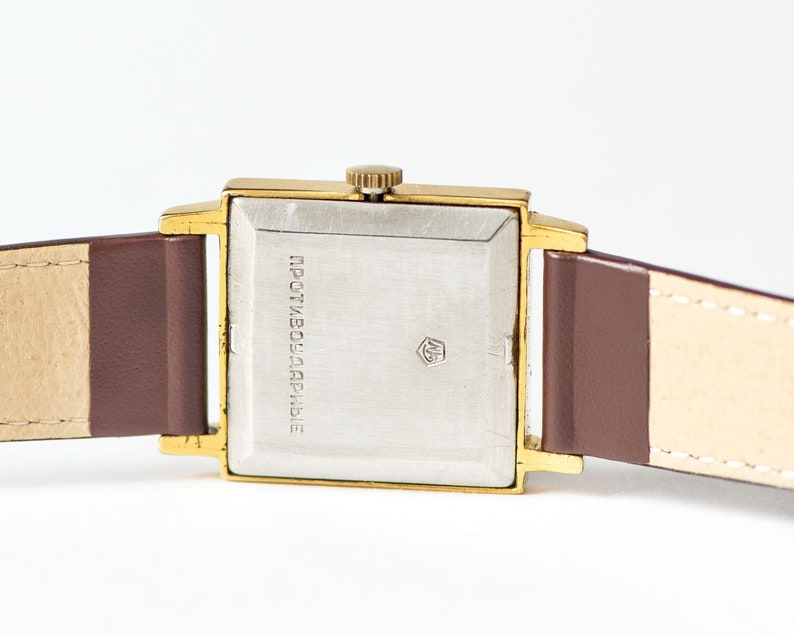 May include: A gold-tone square wristwatch with a brown leather band. The watch face is silver and has the text "ПРОТИВОУДАРНЫЕ" printed on it.