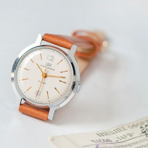 May include: A silver wristwatch with a white face and gold hands. The watch has a brown leather band and the words "Zlatoust" and "12" are printed on the face.