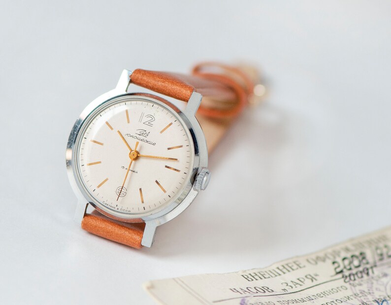 May include: A silver wristwatch with a white face and gold hands. The watch has a brown leather band and the words "Zlatoust" and "12" are printed on the face.