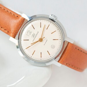 Thin, gold-toned baton stick indexes for the other hours, complemented by matching slim baton-style hands. A round, polished silver-tone case, typically made of chrome-plated brass. Dial: A clean, beige or cream-colored dial featuring a large, stylized Arabic numeral at the 12 o'clock position.