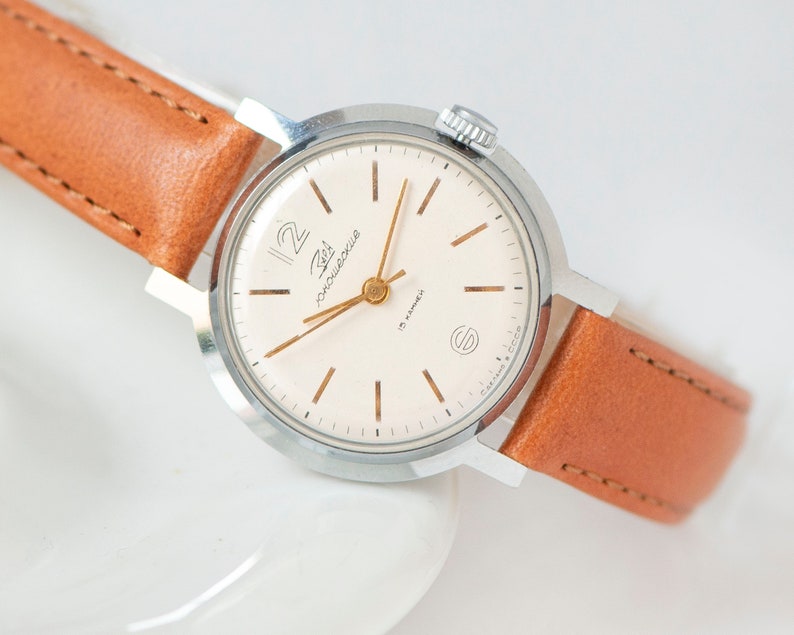 Thin, gold-toned baton stick indexes for the other hours, complemented by matching slim baton-style hands. A round, polished silver-tone case, typically made of chrome-plated brass. Dial: A clean, beige or cream-colored dial featuring a large, stylized Arabic numeral at the 12 o'clock position.