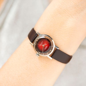 May include: A silver wristwatch with a red face and a brown leather band. The watch face has the text "Automatic 17 Jewels" and "Cristal" printed on it.