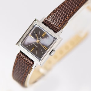 Puede incluir: Un reloj de pulsera cuadrado plateado con una correa de cuero marrón. La esfera del reloj es negra con detalles dorados y el texto "17 камней" en escritura cirílica.