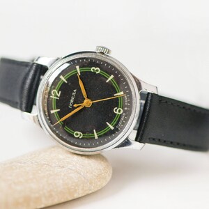 May include: A black and green wristwatch with a silver case and a black leather band. The watch face has white numerals and a gold-colored second hand. The watch face also has the word "Победа" in Cyrillic script, which translates to "Victory" in English.