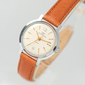 May include: A silver wristwatch with a white face and gold hands. The watch has a brown leather band and says "Zlatoust" on the face.