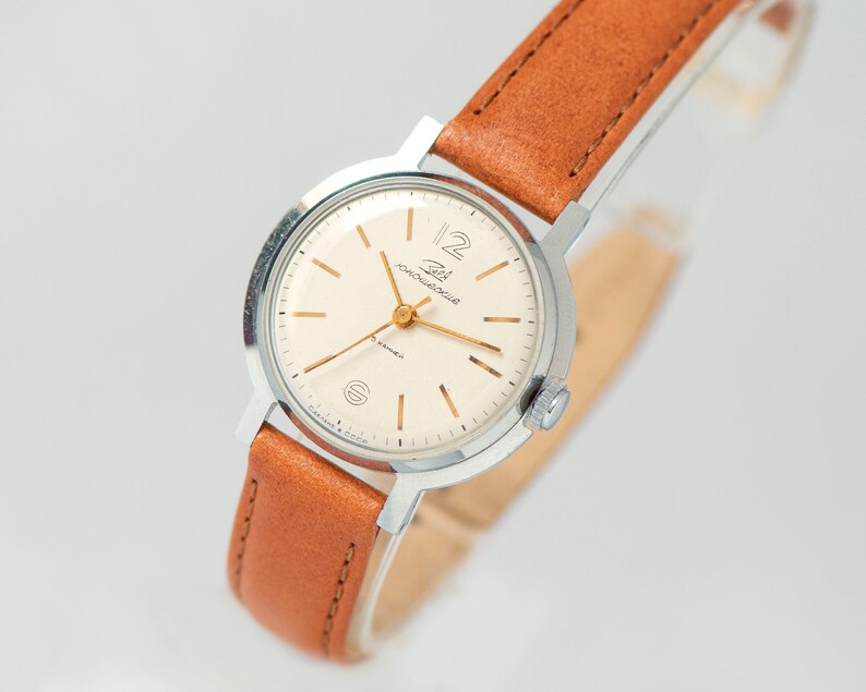 May include: A silver wristwatch with a white face and gold hands. The watch has a brown leather band and says "Zlatoust" on the face.