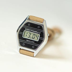 May include: A vintage digital wristwatch with a silver-toned hexagonal case and a tan strap. The black watch face features the text "ЭЛЕКТРОНИКА 5" and "СДЕЛАНО В СССР" with a digital display showing "13.04" and four small circular accents.