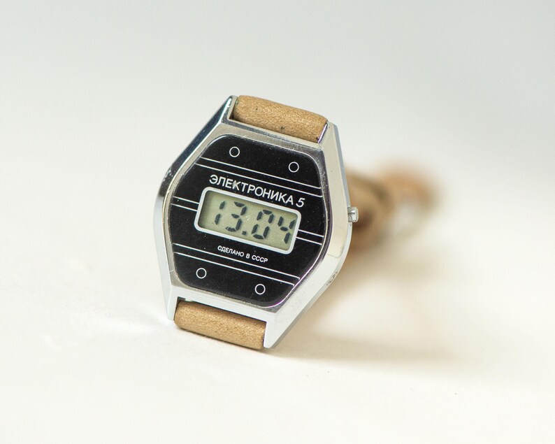 May include: A vintage digital wristwatch with a silver-toned hexagonal case and a tan strap. The black watch face features the text "ЭЛЕКТРОНИКА 5" and "СДЕЛАНО В СССР" with a digital display showing "13.04" and four small circular accents.
