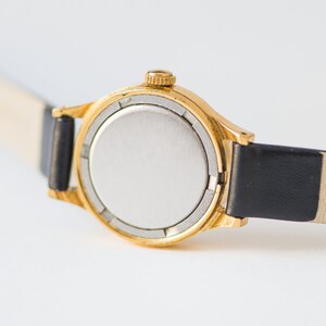 Peut inclure: Une montre-bracelet dor&eacute;e dot&eacute;e d'un bo&icirc;tier rond avec un fond argent&eacute; uni et une petite couronne de remontage sur le c&ocirc;t&eacute;. La montre est fix&eacute;e &agrave; un bracelet en cuir noir fin et lisse qui s'&eacute;tend de chaque c&ocirc;t&eacute; du bo&icirc;tier de la montre.