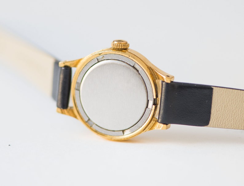 Peut inclure: Une montre-bracelet dor&eacute;e dot&eacute;e d'un bo&icirc;tier rond avec un fond argent&eacute; uni et une petite couronne de remontage sur le c&ocirc;t&eacute;. La montre est fix&eacute;e &agrave; un bracelet en cuir noir fin et lisse qui s'&eacute;tend de chaque c&ocirc;t&eacute; du bo&icirc;tier de la montre.