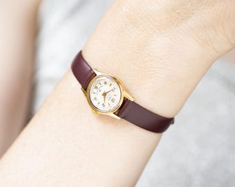 Peut inclure: Une petite montre-bracelet dotée d'un boîtier rond doré et d'un cadran crème avec des chiffres et des aiguilles dorés. La montre est fixée à un bracelet en cuir lisse bordeaux foncé avec une fermeture à boucle simple et une finition polie.