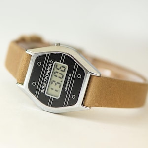 May include: A digital wristwatch with a silver-tone metal case and a tan strap. The black watch face features the text ELEKTRONIKA 5 and СДЕЛАНО В СССР, with a digital display showing the time 13:06. The design includes four small circular accents.