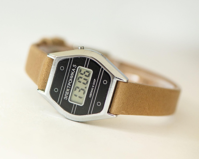 May include: A digital wristwatch with a silver-tone metal case and a tan strap. The black watch face features the text ELEKTRONIKA 5 and СДЕЛАНО В СССР, with a digital display showing the time 13:06. The design includes four small circular accents.