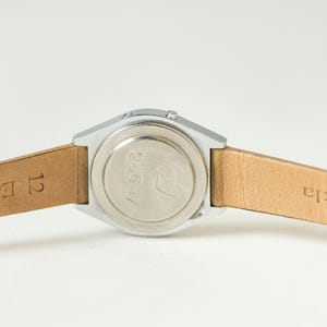 May include: A silver-toned watch case with a circular back plate featuring the engraved numbers 25617 and a small logo. The watch is attached to a light tan leather strap with the text 12 Echt L and Tekla visible on the inner surface.