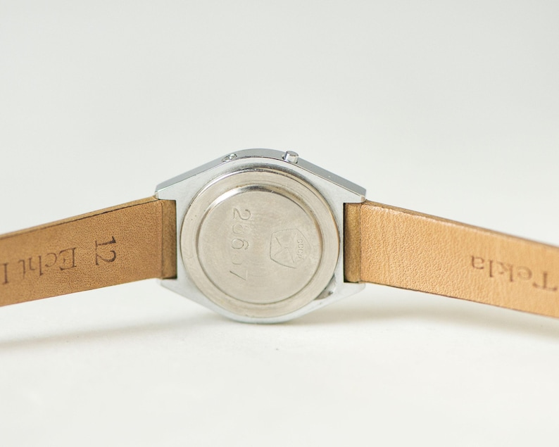 May include: A silver-toned watch case with a circular back plate featuring the engraved numbers 25617 and a small logo. The watch is attached to a light tan leather strap with the text 12 Echt L and Tekla visible on the inner surface.