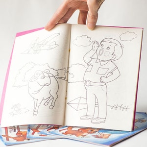 May include: A black and white coloring book with a pink cover. The open page shows a drawing of a boy with his hand to his ear, a sheep, and a plane. The book is on top of other coloring books.