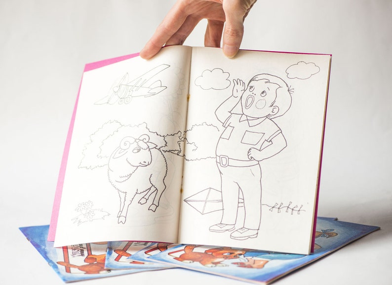 May include: A black and white coloring book with a pink cover. The open page shows a drawing of a boy with his hand to his ear, a sheep, and a plane. The book is on top of other coloring books.