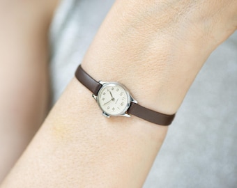 May include: A vintage wristwatch with a silver-colored case and a cream-colored face. The watch has black numerals and hands, and a brown leather band. The watch is shown on a wrist.