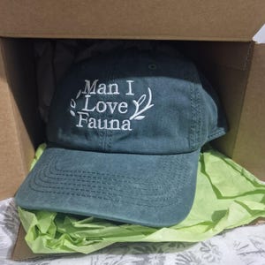 Puede incluir: Gorra de béisbol de color verde azulado con el texto bordado "Man I Love Fauna" en blanco. La gorra está dentro de una caja de cartón, sobre papel de seda verde. El sombrero tiene una visera curva y un diseño cosido.
