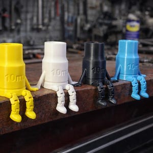 May include: Four colorful, anthropomorphic socket wrenches sit on a metal surface. Each wrench is a different color: yellow, white, black, and blue. The text "10m" is printed on each wrench. They have arms and legs.