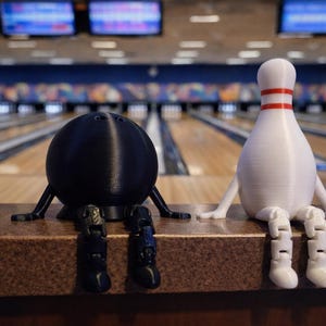 May include: A black bowling ball and a white bowling pin with red stripes, both with arms and legs, sit on a brown surface. The background shows a bowling alley with lanes and screens.