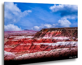 May include: A landscape photograph of a colorful desert canyon under a bright blue sky with fluffy white clouds. The canyon walls display layers of red, white, and brown hues, creating a striking visual contrast. The image is framed with a black border.