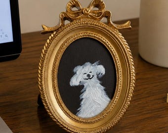 Op de afbeelding: Een ovaalvormige gouden fotolijst met een decoratieve strik aan de bovenkant, met daarin een klein schilderij van een witte hond tegen een zwarte achtergrond. De lijst heeft een getextureerde, touwachtige rand en staat rechtop.
