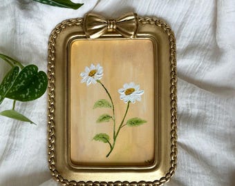 Op de afbeelding: Een kleine, rechthoekige, goudkleurige fotolijst met een strik aan de bovenkant. De lijst bevat een schilderij van twee witte madeliefjes met gele centra en groene stelen tegen een lichtgele achtergrond. De lijst heeft een kralenrand.