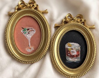 Op de afbeelding: Twee ovale goudkleurige lijsten met strikaccenten aan de bovenkant. De linkerlijst bevat een schilderij van een martiniglas op een perzikkleurige achtergrond. De rechterlijst bevat een schilderij van een cocktail in een glas op een zwarte achtergrond.