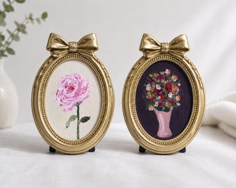 Op de afbeelding: Twee ovale goudkleurige lijsten bekroond met een decoratieve strik. De linkerlijst bevat een schilderij van een enkele roze roos op een lichte achtergrond. De rechterlijst bevat een schilderij van een kleurrijk bloemenboeket in een roze vaas.