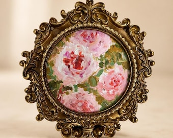 Op de afbeelding: Een rond olieverfschilderij van roze rozen met groen blad, geplaatst in een sierlijke, goudkleurige lijst in antieke stijl. De lijst is voorzien van ingewikkelde krulmotieven en decoratieve reliëfpatronen rondom het ronde bloemenkunstwerk.