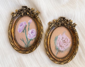 Op de afbeelding: Twee ovale wandlijsten met sierlijke goudkleurige randen met blad- en strikdetails. Elke lijst bevat een klein schilderij op een lichtbruine achtergrond. Eén schilderij toont twee lichtpaarse bloemen, de andere toont een enkele roze roos.